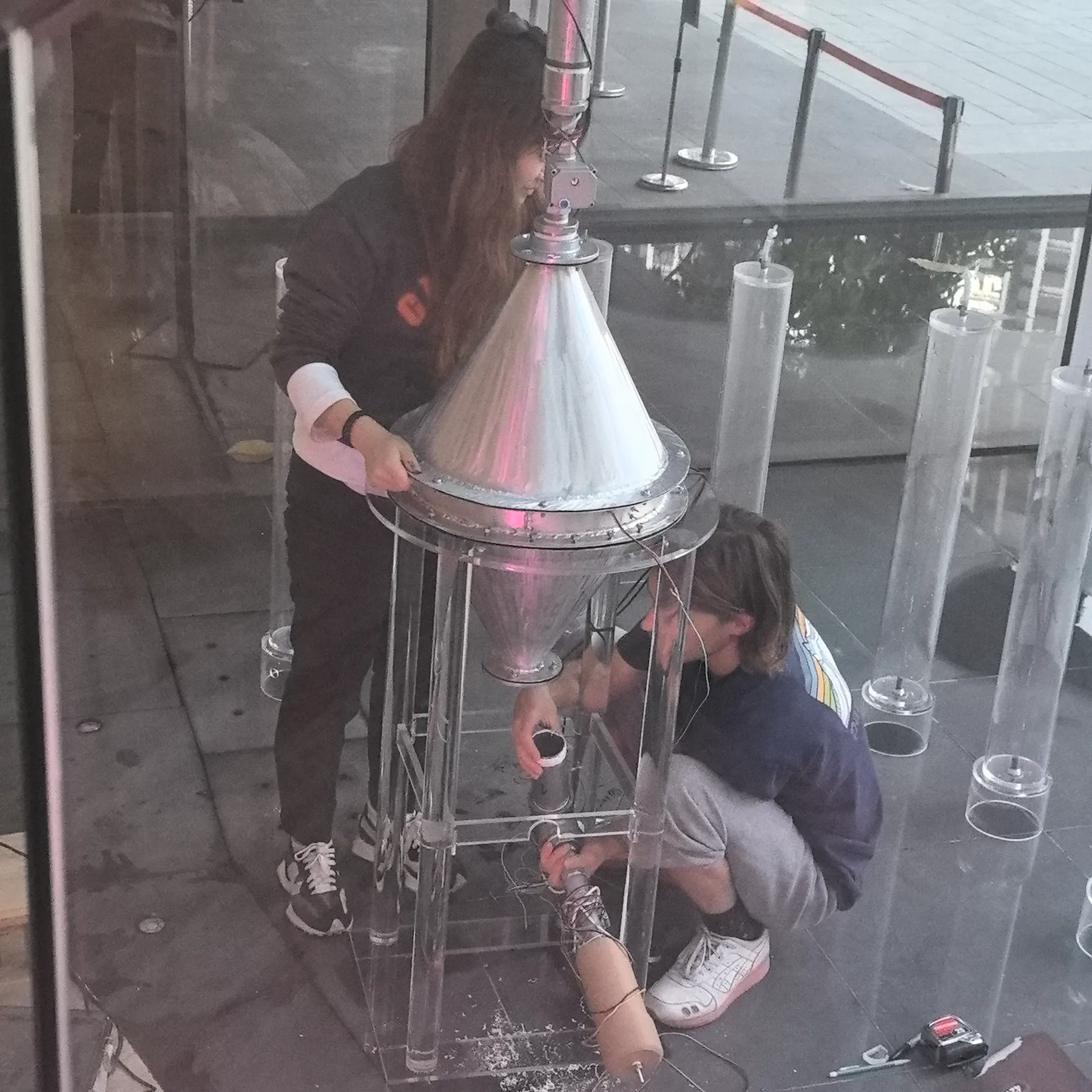 Two team members assembling the steel cone reactor on its clear acrylic frame, with empty algae columns around them.