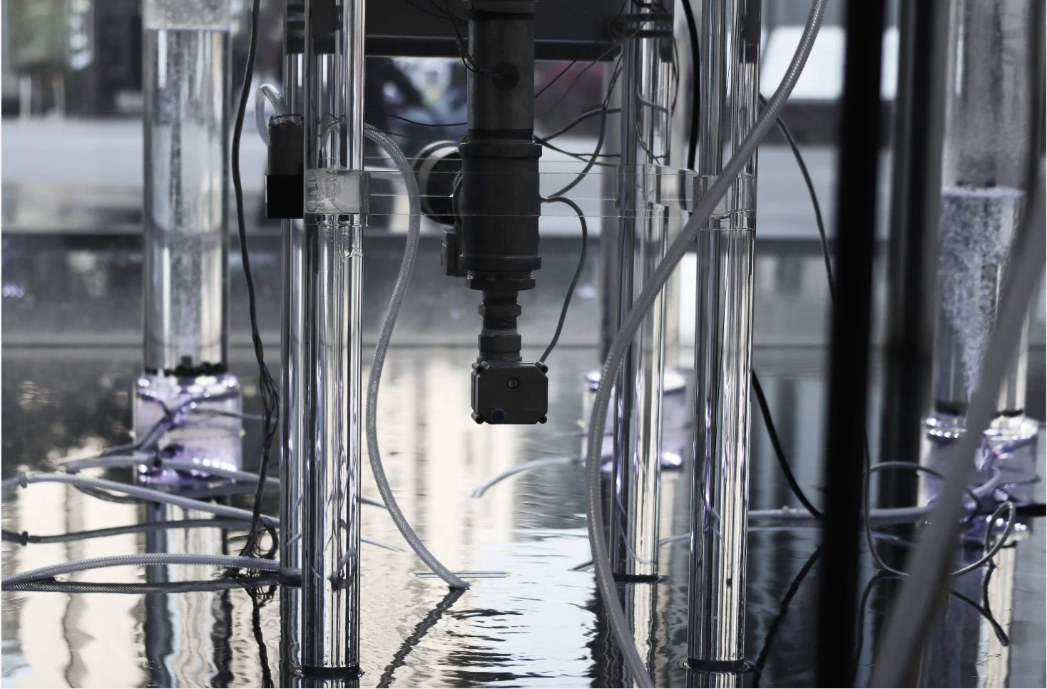 Black and white detail under the reactor: the valve and motor between clear acrylic legs, algae columns bubbling in the background.