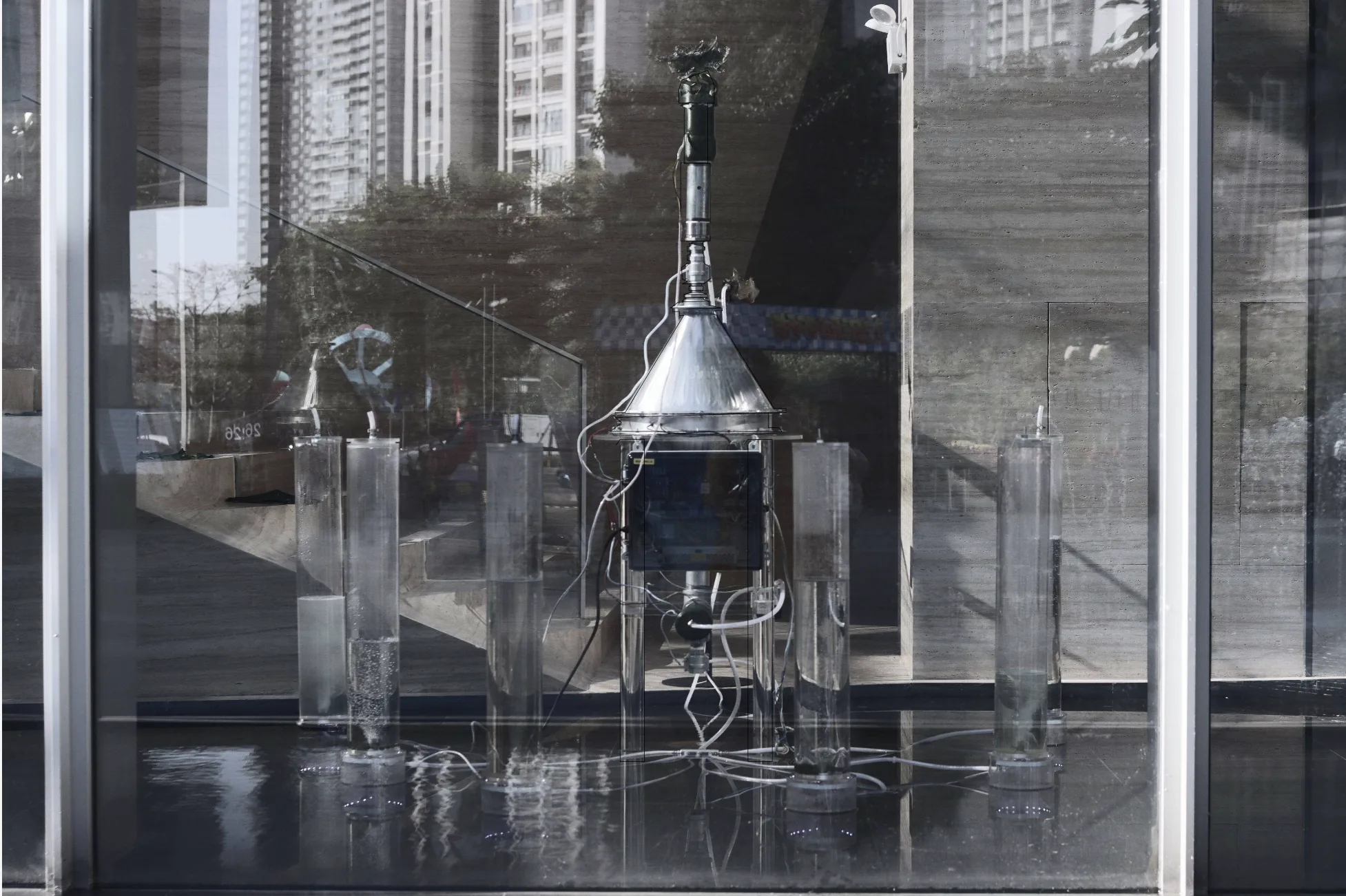 The installation seen from outside through the gallery's glass wall, with the city's apartment towers reflected over the reactor and algae columns.